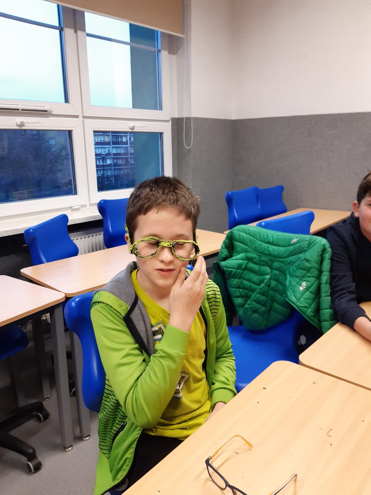 Chłopiec pozuje do zdjęcia w sali lekcyjnej z wykonanymi własnoręcznie okularami z filamentu do długopisów 3D.