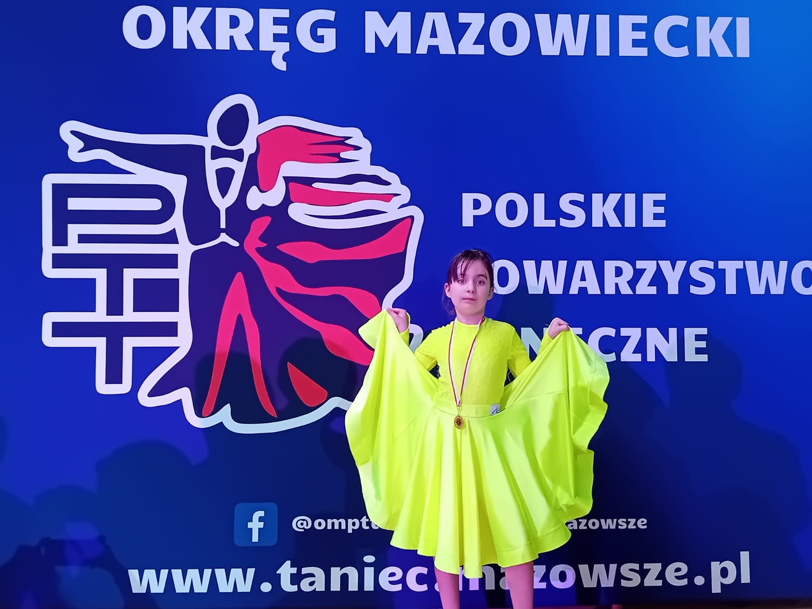 Prezentacja uczennicy w żółtej sukience, w tle baner reklamujący Polskie Towarzystwo Taneczne okręgu mazowieckiego