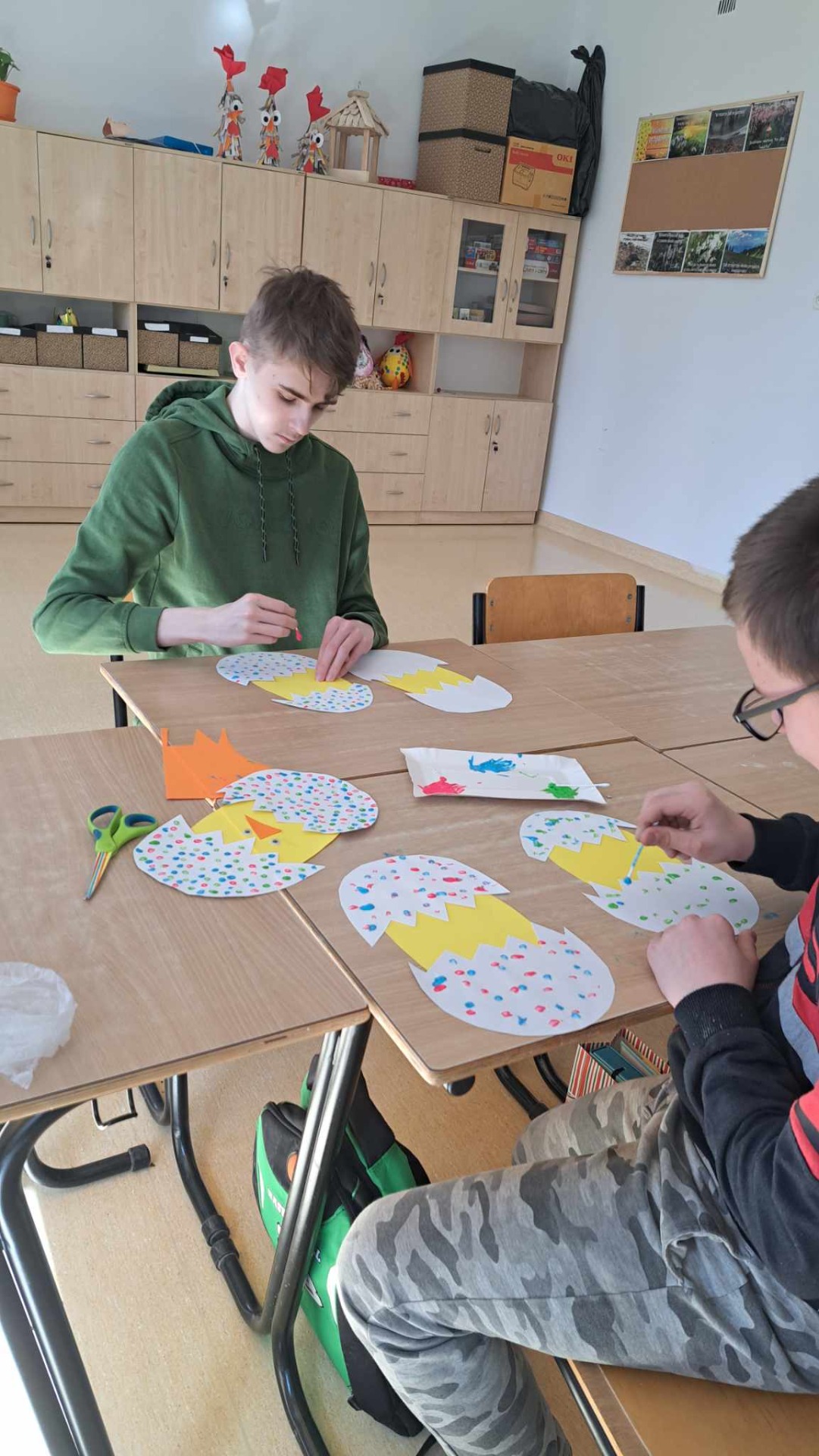 2 uczniów siedzi przy stoliku naprzeciwko siebie - ozdabiają farbami metodą kropkową papierowe łupinki od jajek