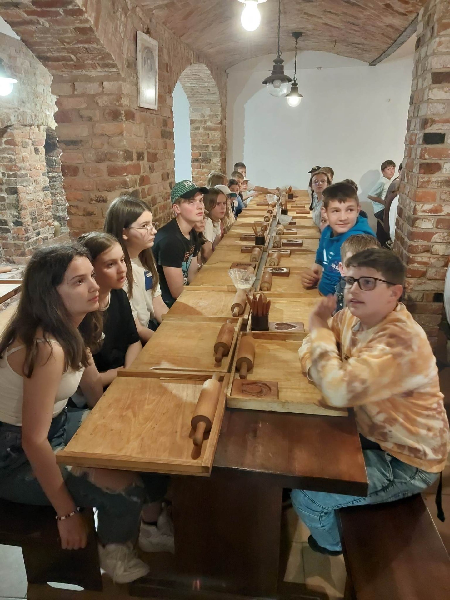 Grupa uczniów siedzi przy drewnianym stole w czasie warsztatów wyrabiani piernika