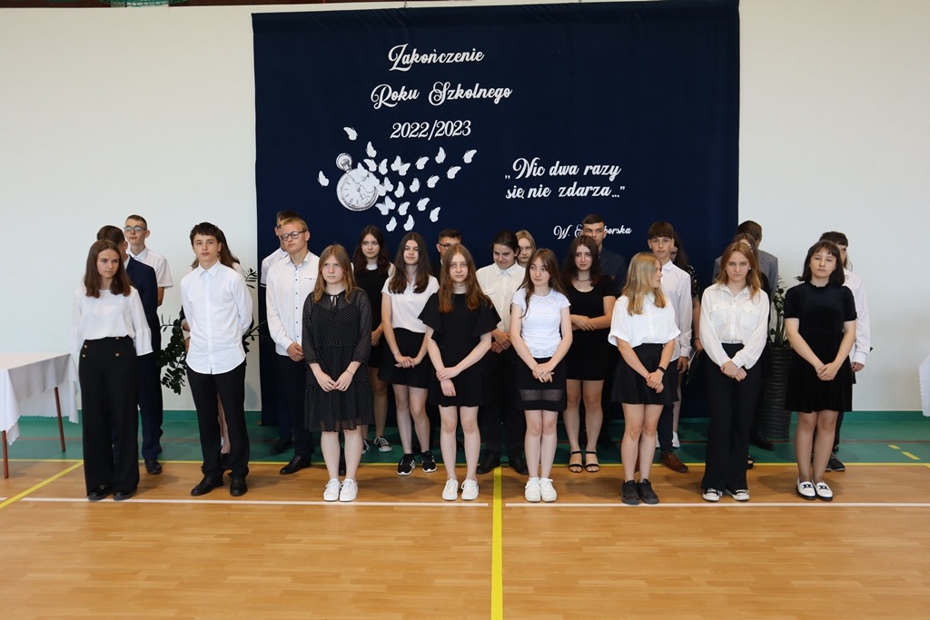 Fotografia przedstawia uczniów klasy ósmej a oraz klasy ósmej b stojących na sali gimnastycznej. W tle okolicznościowa dekoracja.
