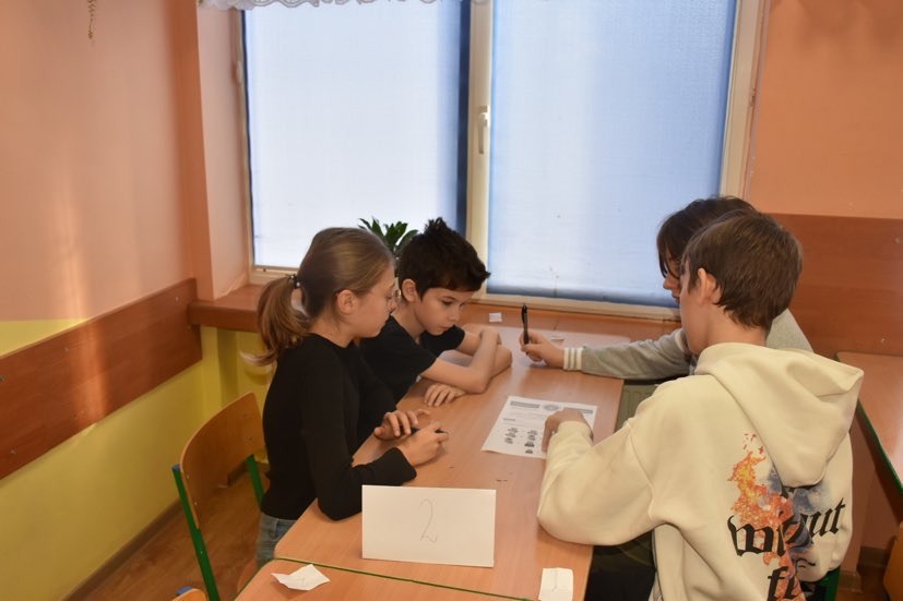 Uczniowie siedzą przy stolikach i w grupach rozwiązują zadania konkursowe