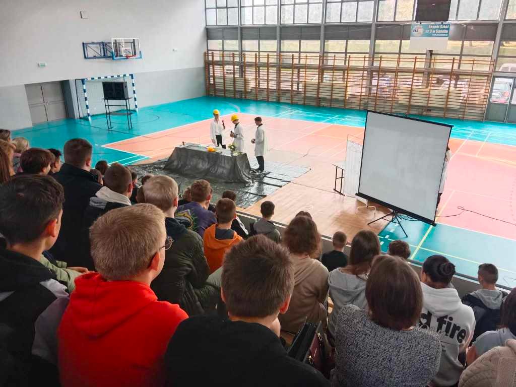 Na zdjęciu widać sale gimnastyczna i uczniów siedzących na trybunach, oglądających pokaz z fizyki i chemii.