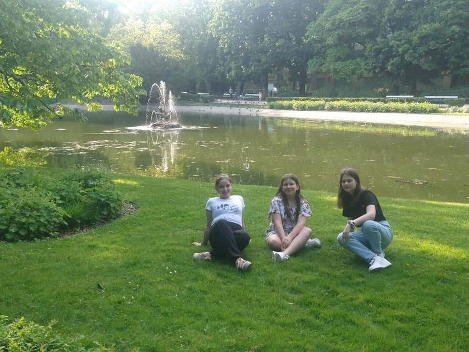 Zdjęcie przedstawia trzy uczennice siedzące na trawie w Ogrodzie Saskim. W tle drzewa, krzewy, jezioro, w środku którego znajduje się fontanna. 