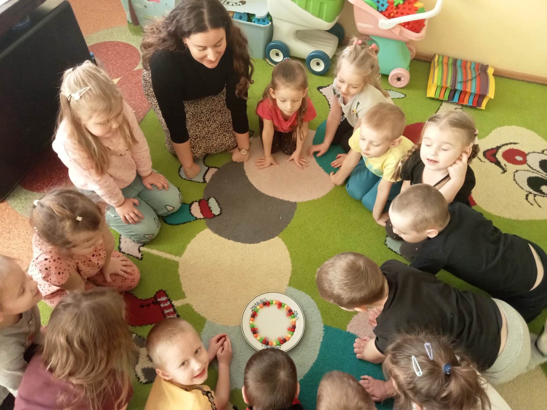 Dzieci siedzą w kole na kolorowym dywanie w sali przedszkolnej i biorą udział w warsztatach w ramach projektu edukacyjnego "Gramy zmysłami"