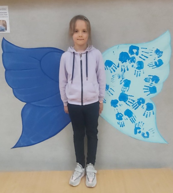 Dziewczynka stoi plecami do niebieskich skrzydeł powieszonych na ścianie.