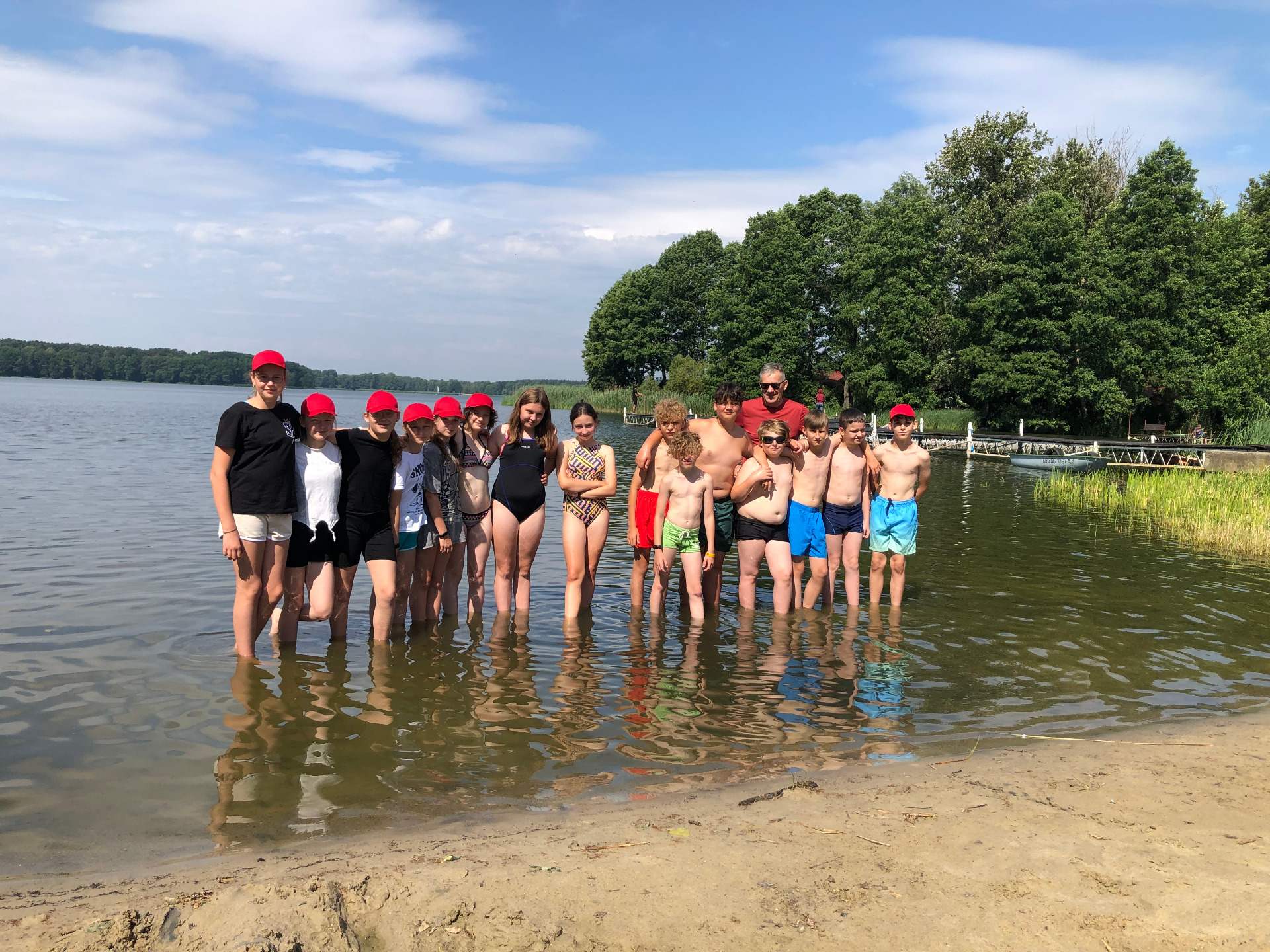 Fotografia przedstawia grupę  uczestników obozu rekreacyjno-sportowego z opiekunem. Grupa stoi  w wodzie na brzegu plaży w tle jezioro i pomost.