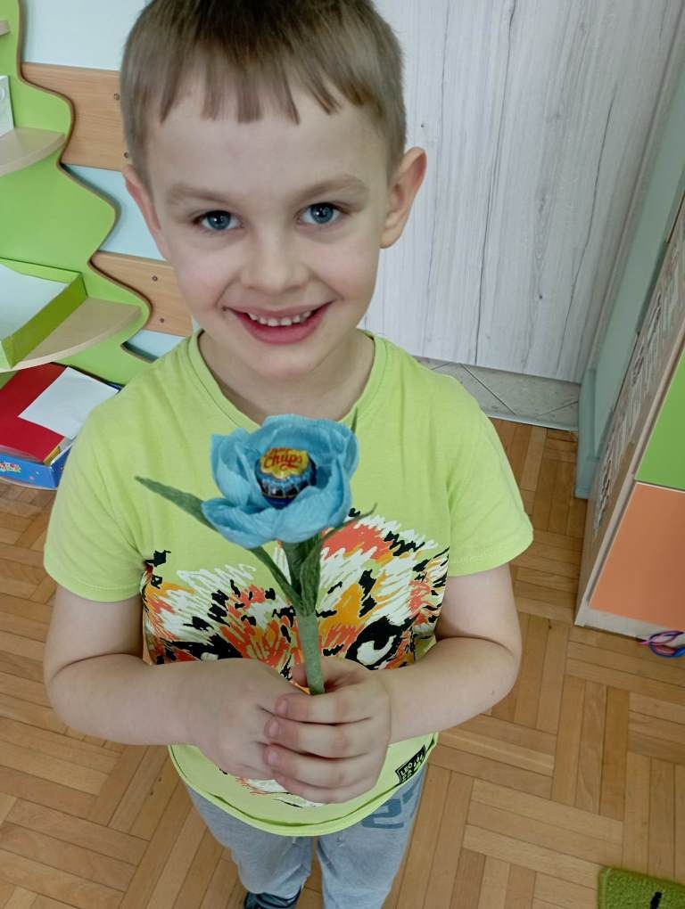 Na piątym zdjęciu stoi uśmiechnięty chłopiec trzymający w ręce niebieski kwiatek. 