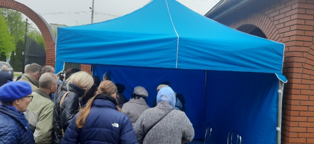 Pod niebieskim namiotem dziewięcioro ludzi odwróconych tyłem.
