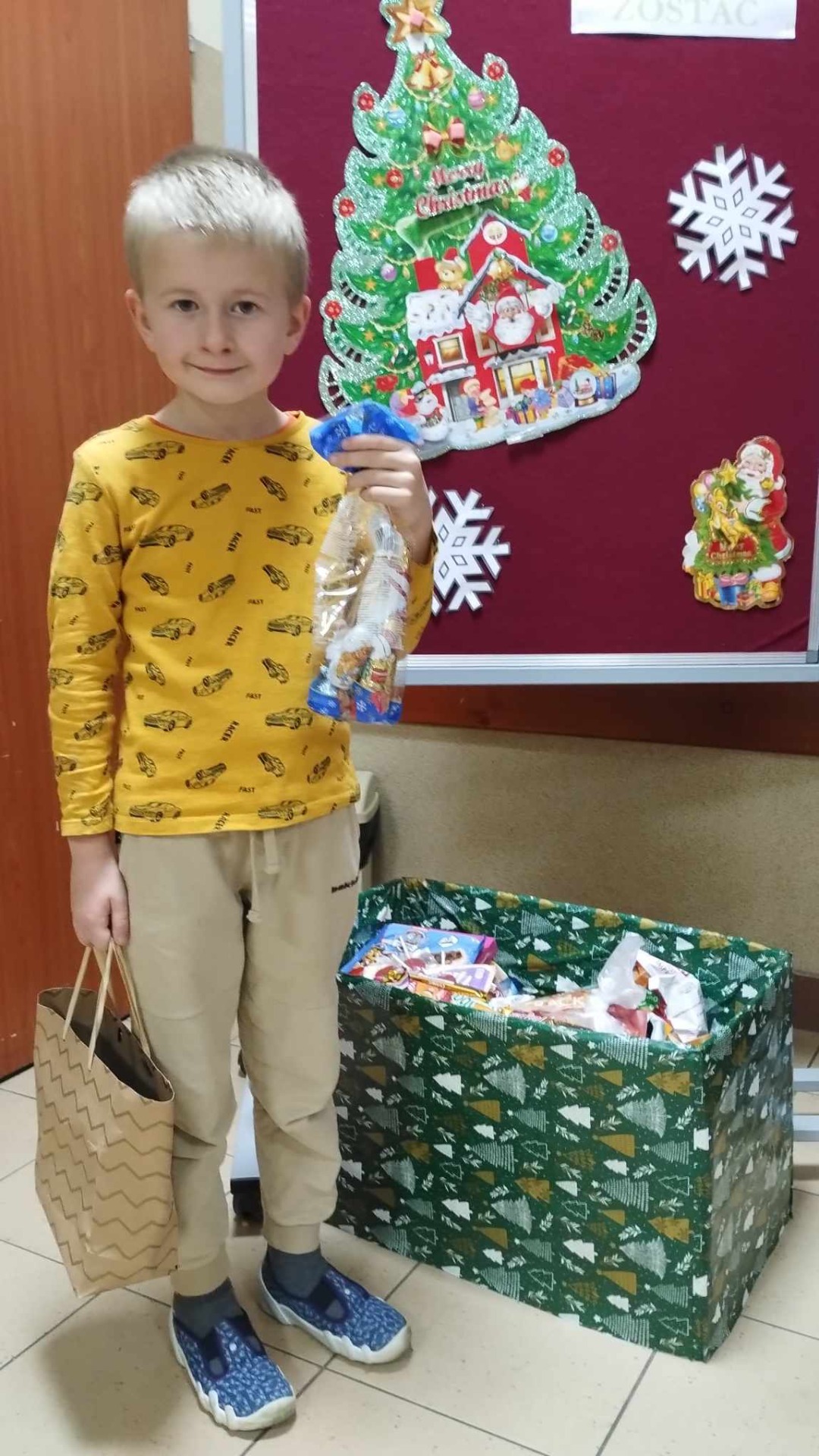Dzieci wrzucają słodkie upominki do pudełka z darami obok gazetki z napisem "I Ty możesz zostać Świętym Mikołajem".