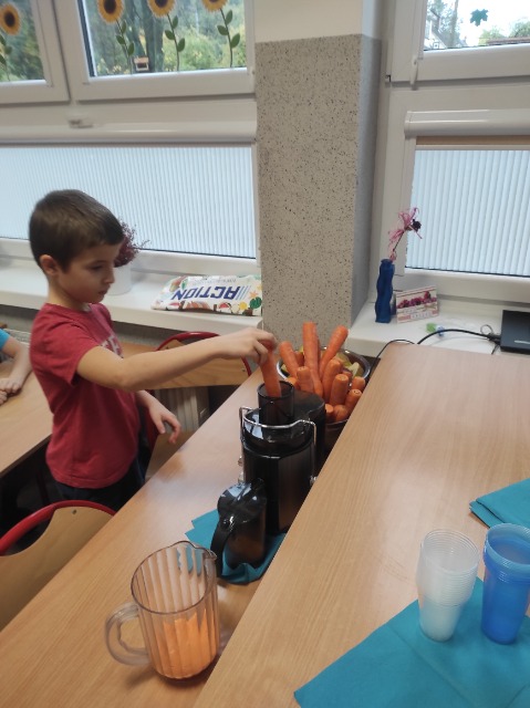 Przed stolikiem stoi chłopiec, który wkłada marchewkę do sokowirówki. Obok widać stojące marchewki i dzbanek z sokiem
