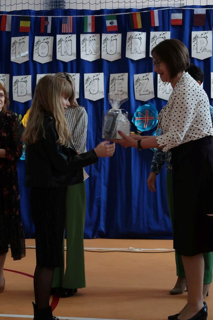 Na zdjęciu po prawej stronie kobieta (Wice Dyrektor Zespołu Szkol w Niebocku - Pani Renata Sieńczak ) wręcza nagrodę dziewczynce o długich jasnych włosach. W tle widać niebieskie płótno z napisem "Konkurs" na białych kartkach.